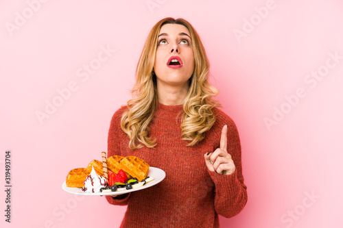 Young blonde woman eating a waffle dessert isolated pointing upside with opened mouth.