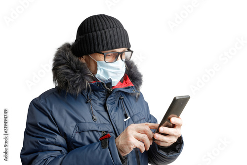 Man in winter clothes, black hat, glasses and a protective mask holds a smartphone in his hands and looks on the screen. Isolated on white.