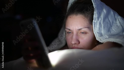 a girl without makeup uses a phone at night, lying under a blanket, she has insomnia