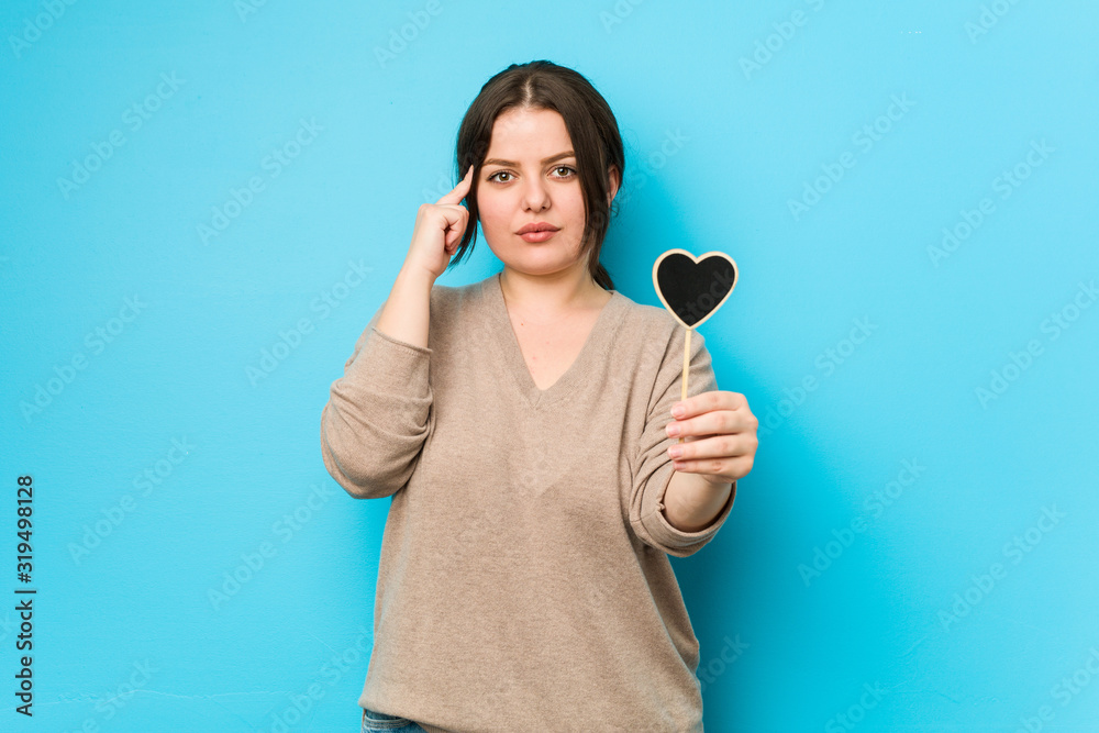 Young plus size curvy woman holding a heart shape pointing his temple with finger, thinking, focused on a task. Young plus size curvy woman holding a heart shape pointing his temple with finger, thinking, focused on a task.