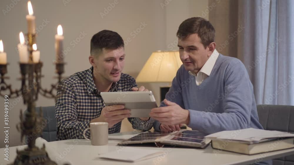Portrait of positive adult Caucasian son teaching middle aged father to use tablet. Man showing parent how to surf Internet. Generations, lifestyle, family. Portrait of positive adult Caucasian son teaching middle aged father to use tablet. Man showing parent how to surf Internet. Generations, lifestyle, family.