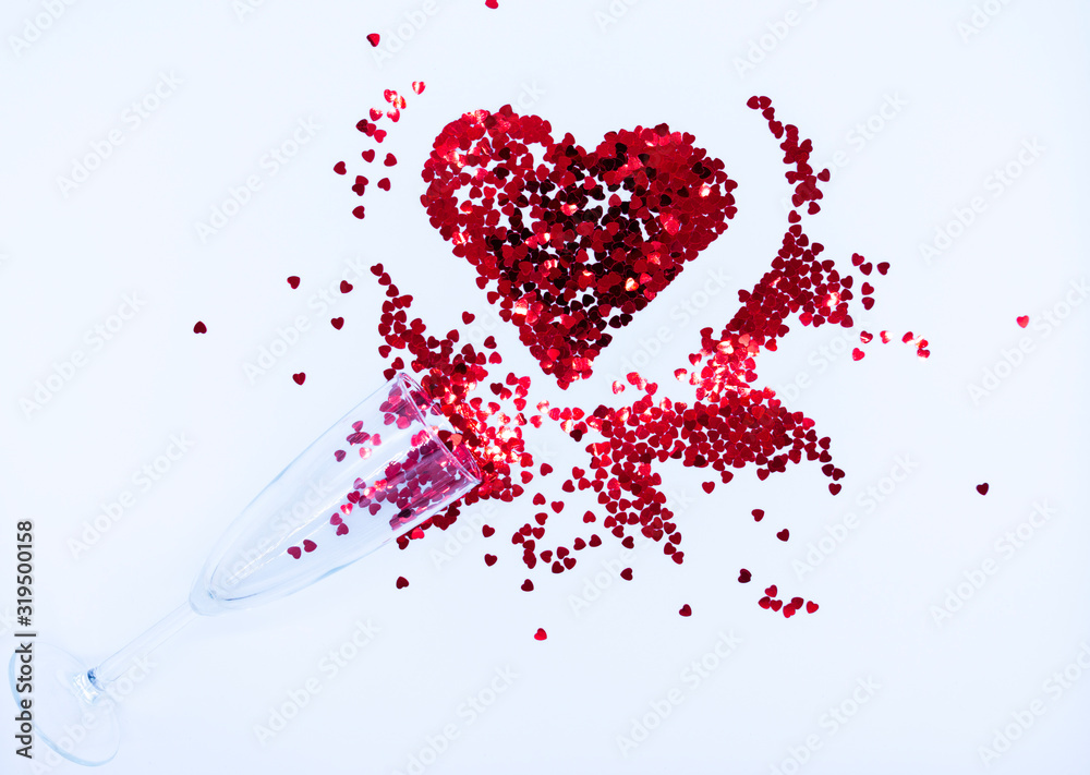 Champagne glass with red sequins scattered in the form of a heart on a white backdrop. Background for Valentine's day. Top view. Flat lay.