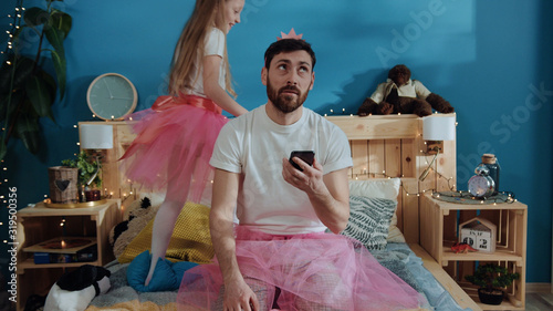 Portrait of tired bored young man in fairy dress using a smartphone sitting on bed. Happy princess girl dancing around waving magic wand playing with her daddy at home. photo