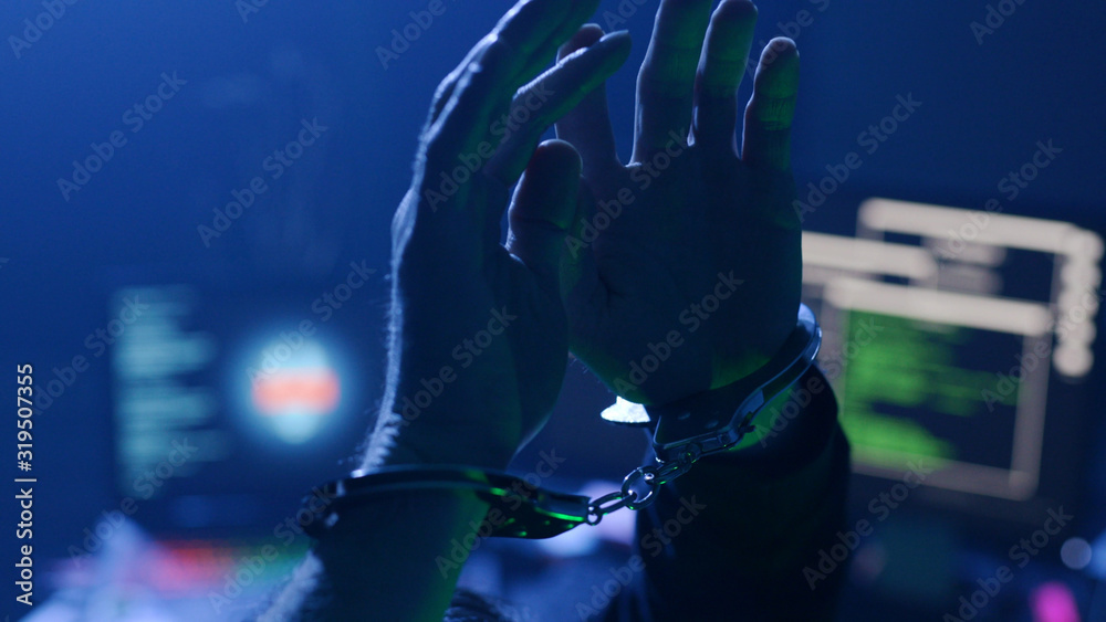 Close-up of male hacker arrested for cyber crime wearing handcuffs. View of computer hacker�s workplace in smoky room on background. Close-up of male hacker arrested for cyber crime wearing handcuffs. View of computer hacker�s workplace in smoky room on background.