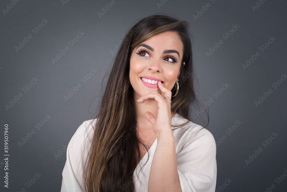 Dreamy female with thoughtful expression, looks to the camera, keeps hand near face, bitting a finger thinks about something pleasant, poses against gray wall. Dreamy female with thoughtful expression, looks to the camera, keeps hand near face, bitting a finger thinks about something pleasant, poses against gray wall.