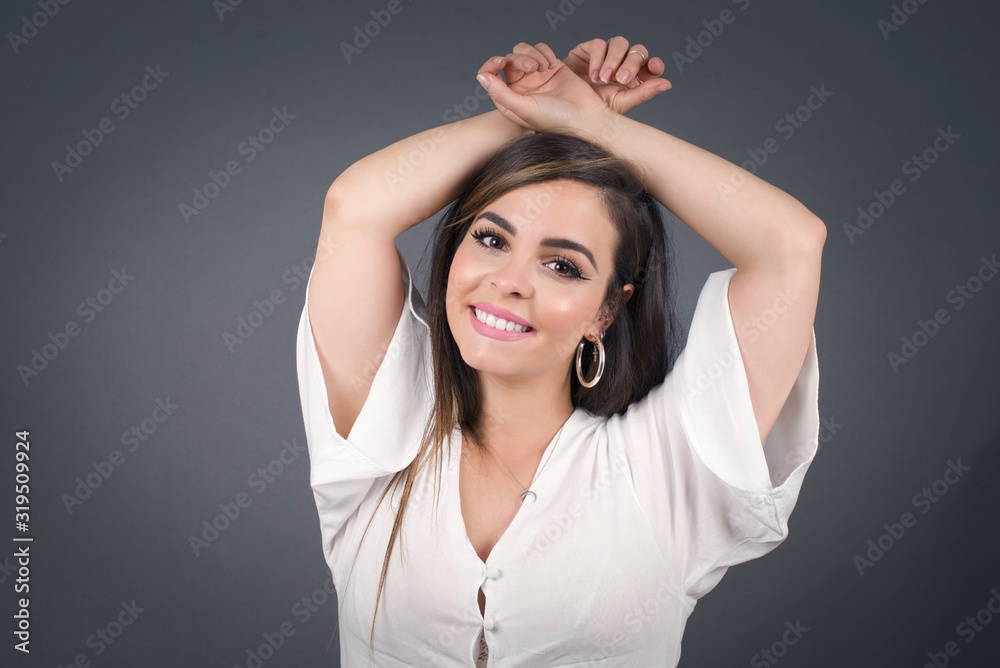 Portrait of beautiful brunette female looks with excitement at camera, keeps hands raised over head notices something unexpected, isolated over gray wall. Lovely woman reacts on sudden news.