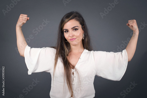 Waist up shot of caucasian woman raises arms to show her muscles feels confident in victory, looks strong and independent, smiles positively at camera, stands against gray background. Sport concept.