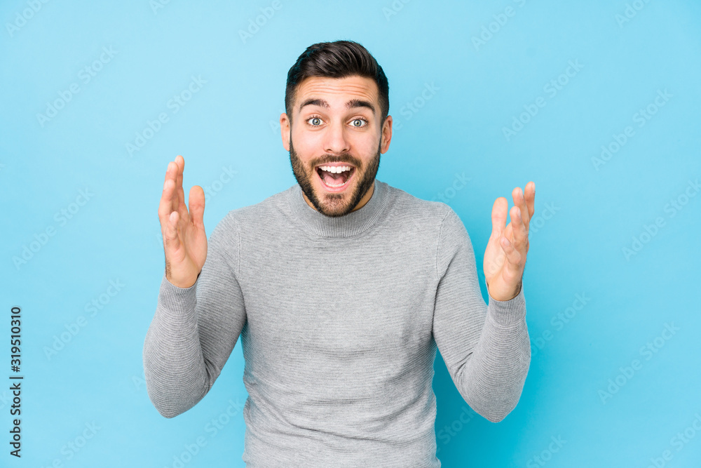 Naklejka premium Young caucasian man against a blue background isolated receiving a pleasant surprise, excited and raising hands.
