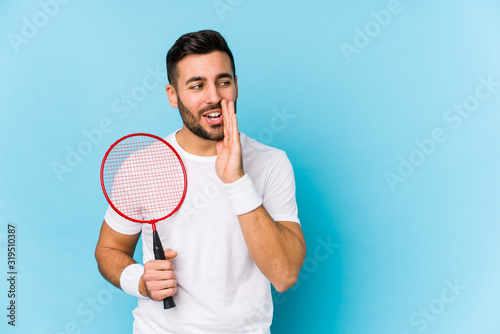 Young handsome man playing badminton isolated is saying a secret hot braking news and looking aside