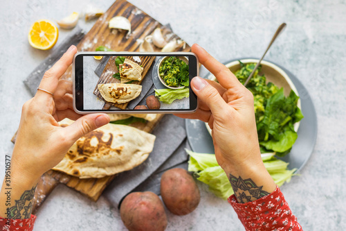 Woman take picture of vegan food with phone at her kitchen. Hand make a closeup smartphone photo of green salad and stuffed bread for blogging or social media content. Vegetarian healthy food.