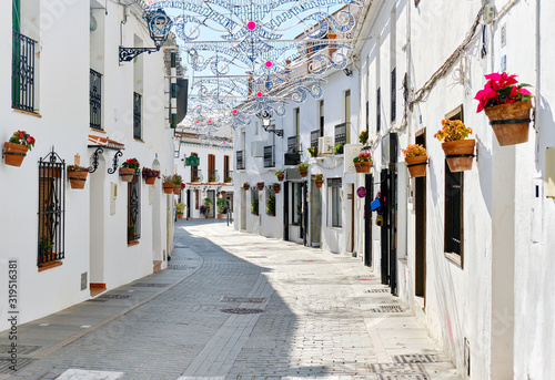 Fototapeta Naklejka Na Ścianę i Meble -  Mijas white washed street, small famous village in Spain. Charming empty narrow streets with New Year decorations, on houses walls hanging flower pots, sunny day no people. Costa del Sol, Málaga