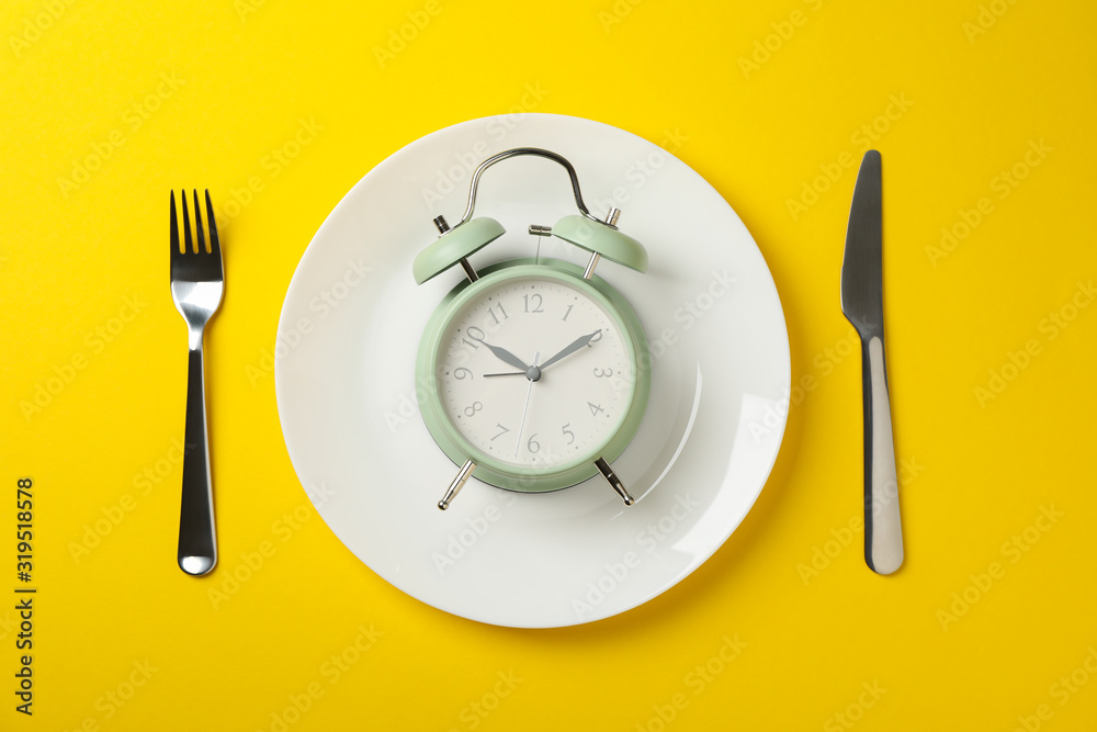 Plate with alarm clock, fork and knife on yellow background, top view Plate with alarm clock, fork and knife on yellow background, top view