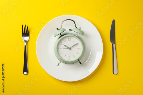 Plate with alarm clock, fork and knife on yellow background, top view