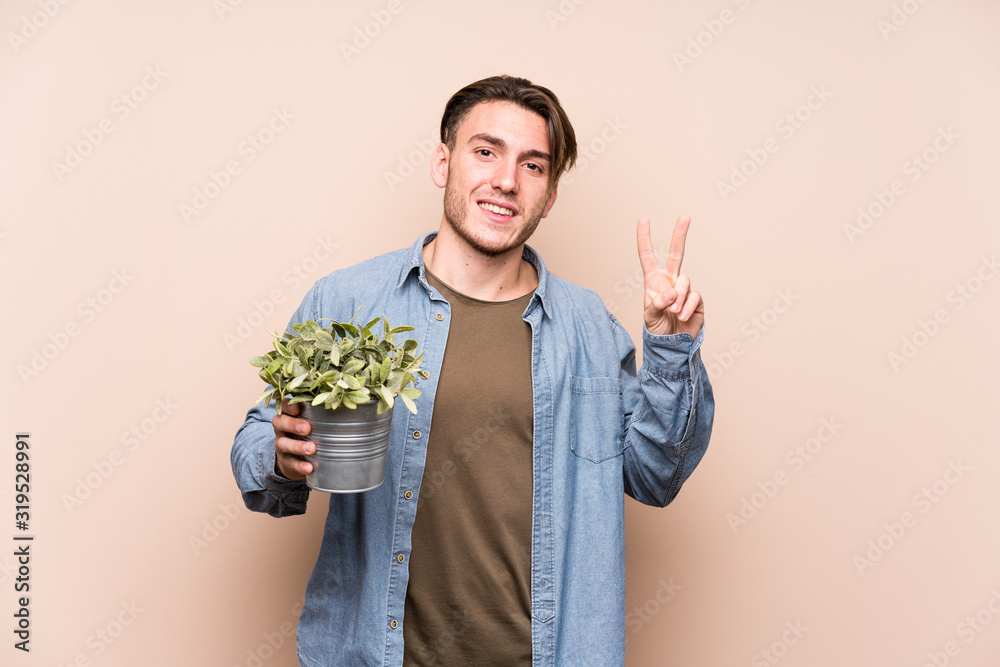 Young caucasian man holding a plant joyful and carefree showing a peace symbol with fingers. Young caucasian man holding a plant joyful and carefree showing a peace symbol with fingers.