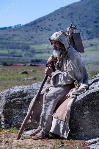 Beggar, ragged, holding on a stick, climbed on a rock in the countryside photo