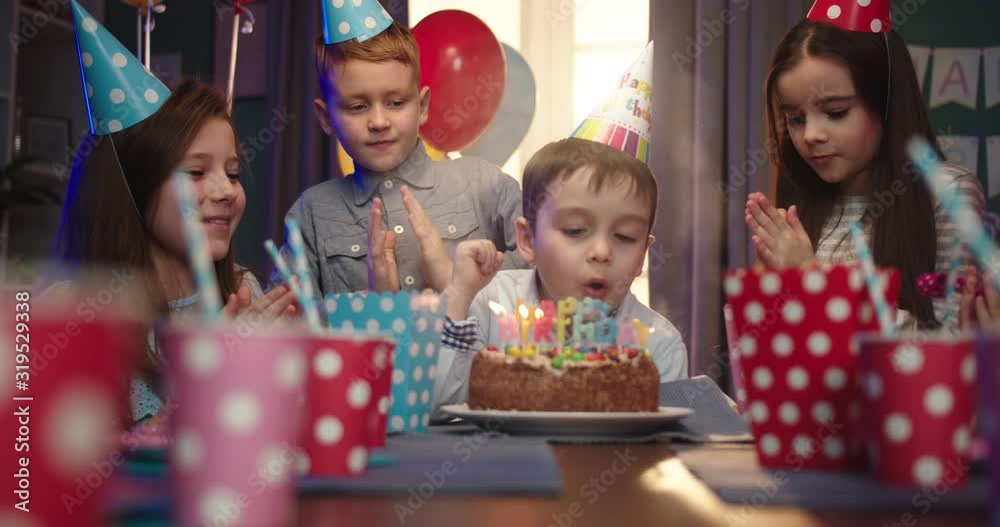 Happy little Caucasian boy celebrating a birthday - blowing candles and smiling at the camera while his friends blowing in horns and applausing. Happy little Caucasian boy celebrating a birthday - blowing candles and smiling at the camera while his friends blowing in horns and applausing.