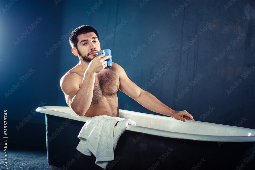 Handsome naked muscular adult bearded man taking bath and drinking coffee or tea, isolated on dark-blue background (with copyspace for your text) - obrazy, fototapety, plakaty 
