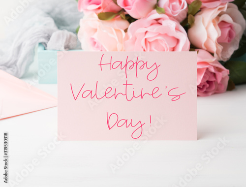 Happy Valentines Day written on pink paper card with rose flowers in the background with envelope