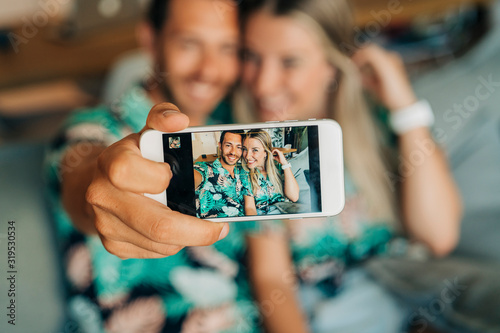 Happy couple sitting on couch in living room wearing Hawaiian shirts taking a selfie photo