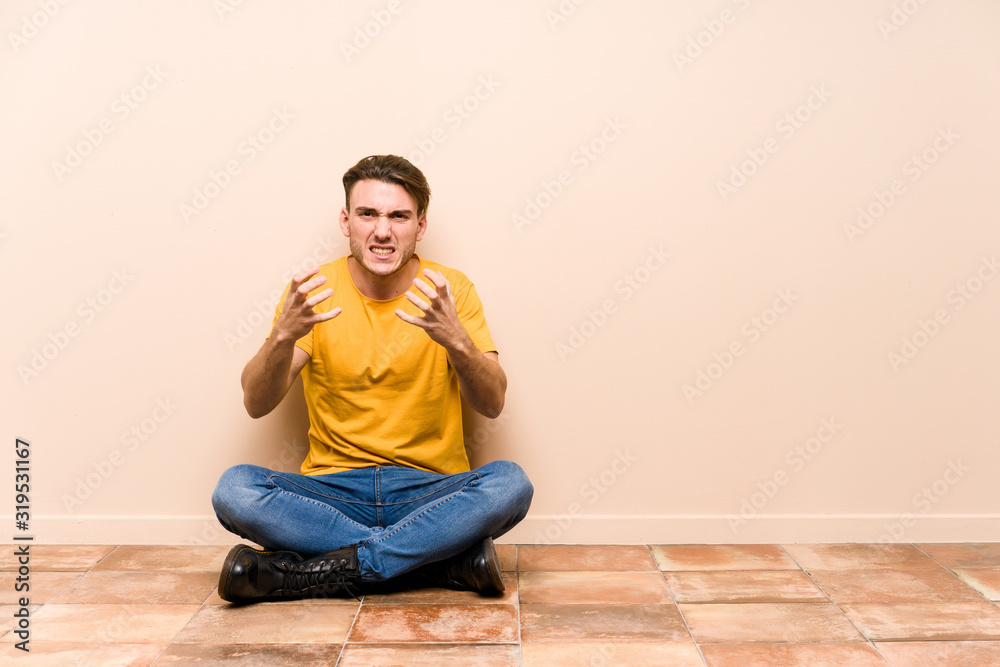 Young caucasian man sitting on the floor isolated upset screaming with tense hands.