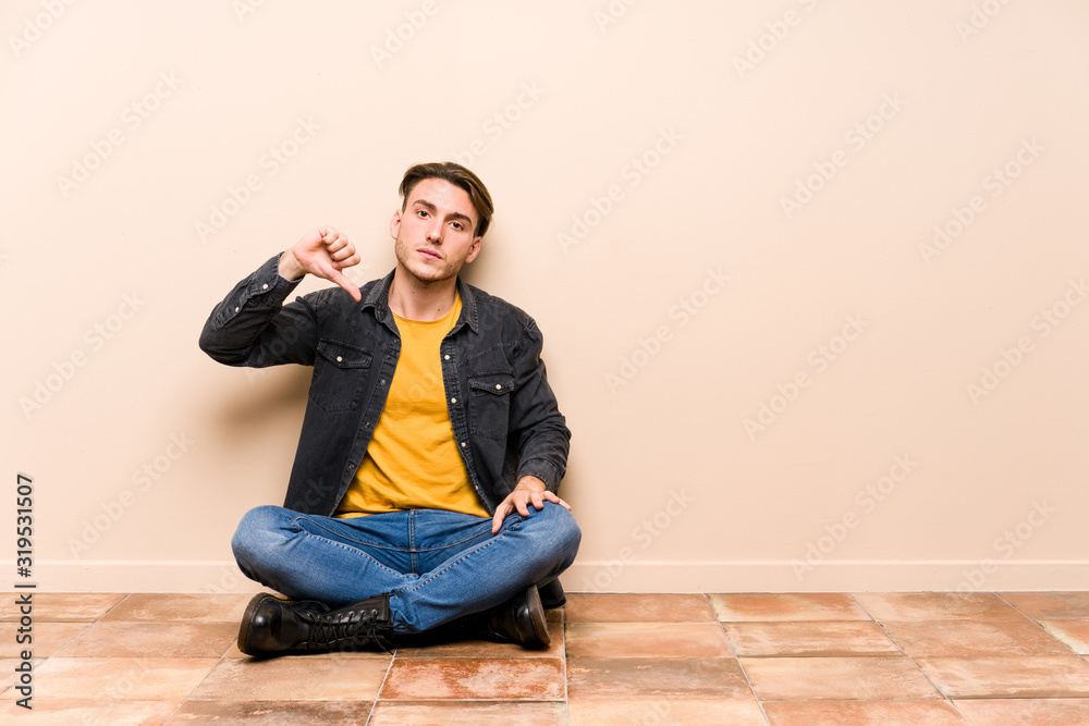 Young caucasian man sitting on the floor isolated showing a dislike gesture, thumbs down. Disagreement concept.