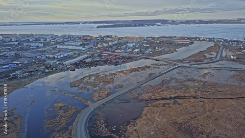 pictorial town panorama with small buildings surrounded by calm river water under sky with clouds in spring tundra aerial