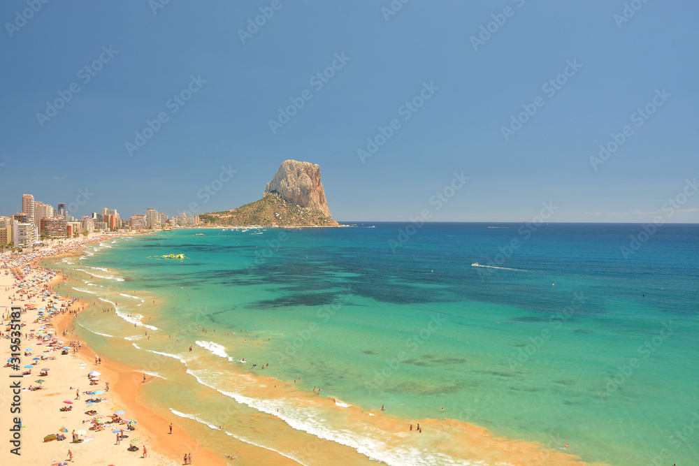 Colorful Mediterranean seascape.People sunbathe on the on the sea coast under umbrellas. In the distance, high-rise buildings and hotels of the resort town of Calpe and Mountain Penyal d'Ifach. Spain.