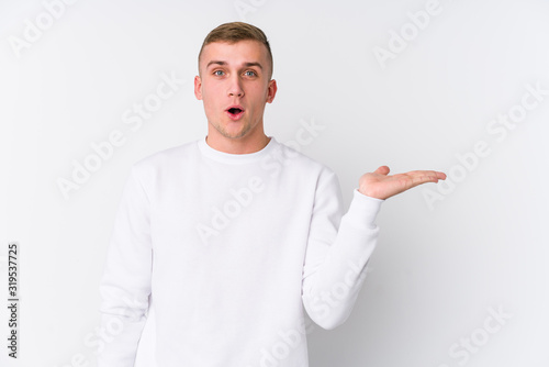 Young caucasian man on white background impressed holding copy space on palm.