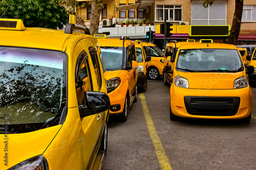 Group of yellow taxis parked in the city center under the trees. On the roofs and sides of cars there are colorful reflections, shadows and sun glare. Selective focus