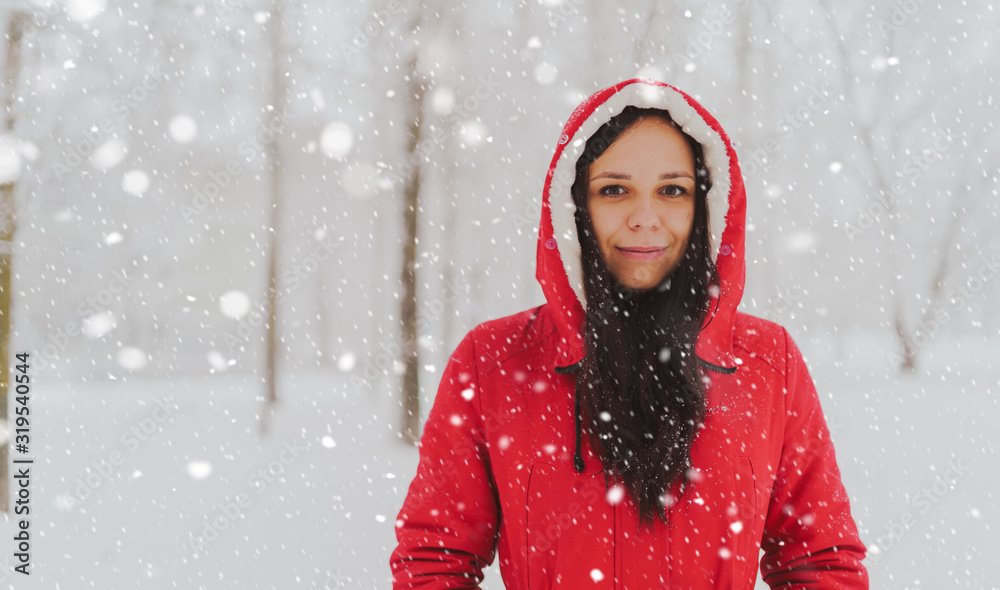 Portrait of young woman in red jacket and hood in winter season. Beautiful lady smiles, rejoicing in fall of snow. Fluffy snow envelops everything around.
