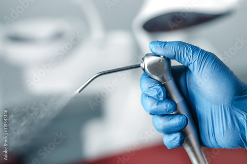Female hand of dentist in blue gloves holding air water syringe, blurred background of clinic with dental unit, dentistry and teeth care concept with copy space