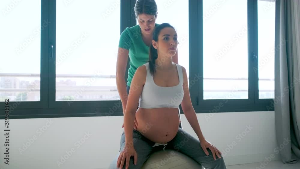 Video of physiotherapist helping to beautiful pregnant woman for doing pilates exercises with ball preparing for childbirth.