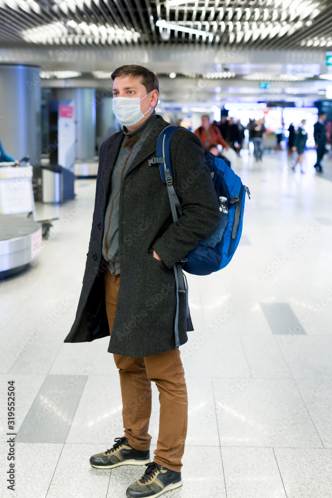 Young European man in gray coat, protective disposable medical mask in airport. Afraid of dangerous N-CoV 2019 influenza coronavirus mutated and spreading in China. Blue backpack, suitcase on wheels Young European man in gray coat, protective disposable medical mask in airport. Afraid of dangerous N-CoV 2019 influenza coronavirus mutated and spreading in China. Blue backpack, suitcase on wheels
