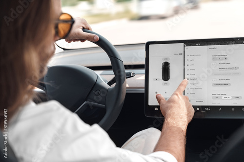 Mode of Transport. Bearded man with long hair in sunglasses traveling sitting inside electric car driving choosing autopilot on smart system close-up photo