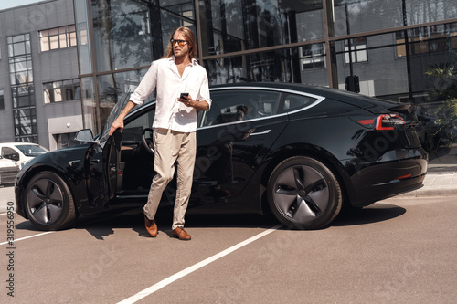 Mode of Transport. Man with long hair going out of electric car with smartphone looking aside curious © Viktoriia