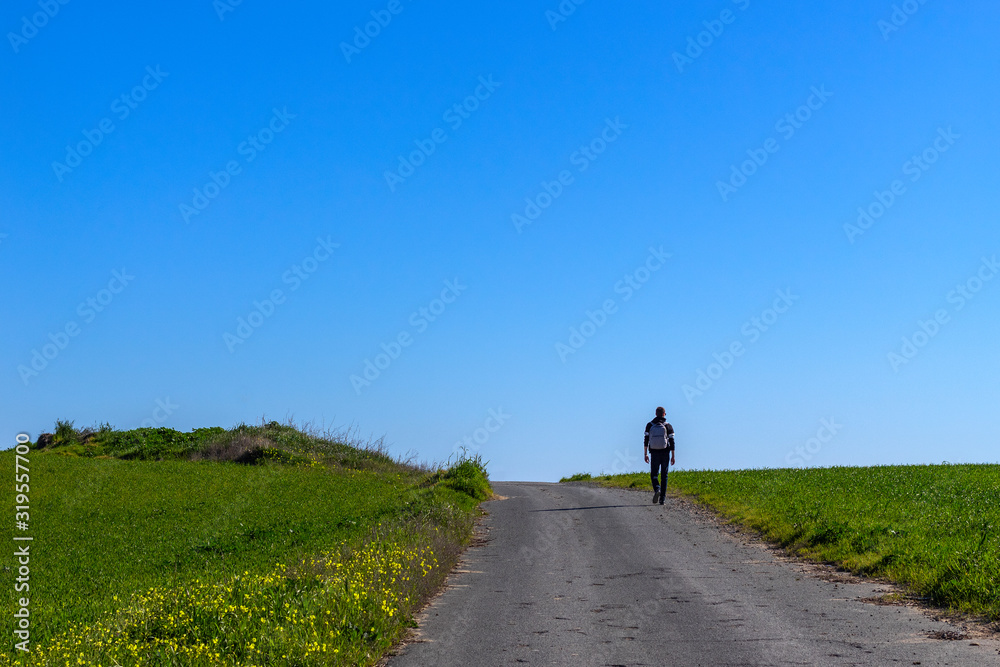 Obraz premium A lonely backpacker tourist walks the road through a large field covered with green grass.