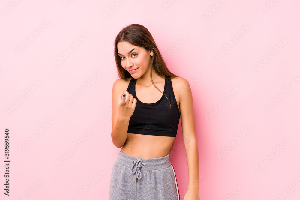 Young caucasian fitness woman posing in a pink background pointing with finger at you as if inviting come closer.