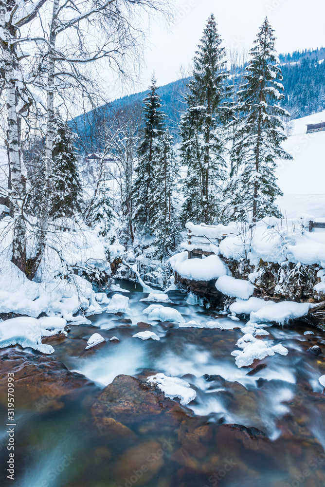 Winter landscape of a small mountain river with colorful stones and frozen waterfall in Alps. The trees in the forest and the stones in stream are covered with snow. Winter landscape of a small mountain river with colorful stones and frozen waterfall in Alps. The trees in the forest and the stones in stream are covered with snow.
