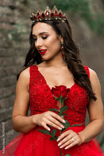 Beautiful brunette in a red dress with a crown. A brunette girl with one red rose outside on stone loft wall background. Artistic photo. Wedding photosoot photo