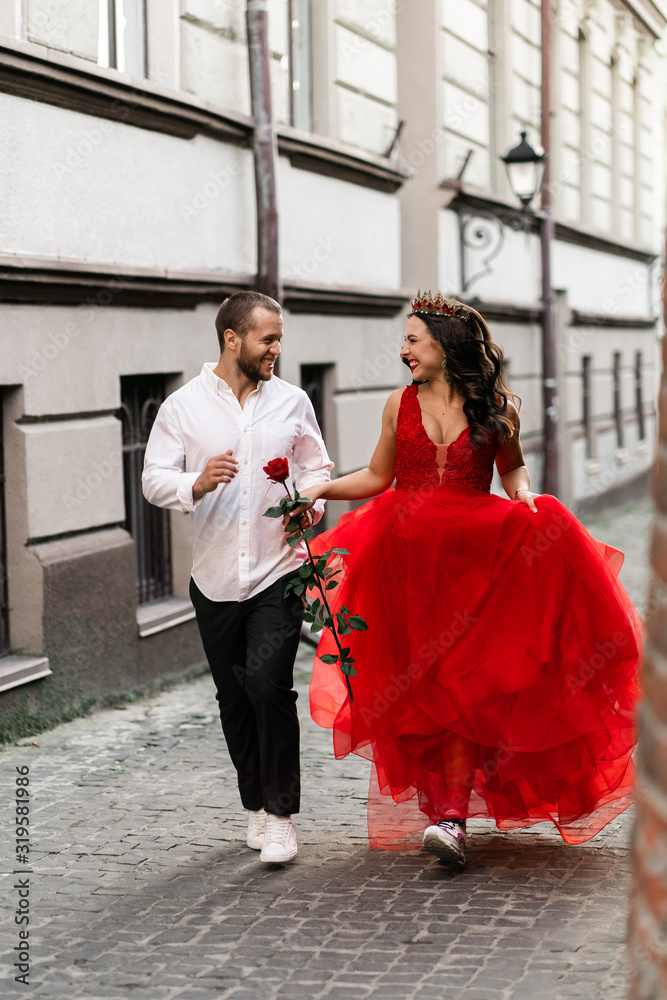 Beautiful romantic couple. Attractive young woman in red dress and crown with handsome man in white shirt with red rose walking on the street.Happy Saint Valentine's Day. Pregnant and wedding concept.