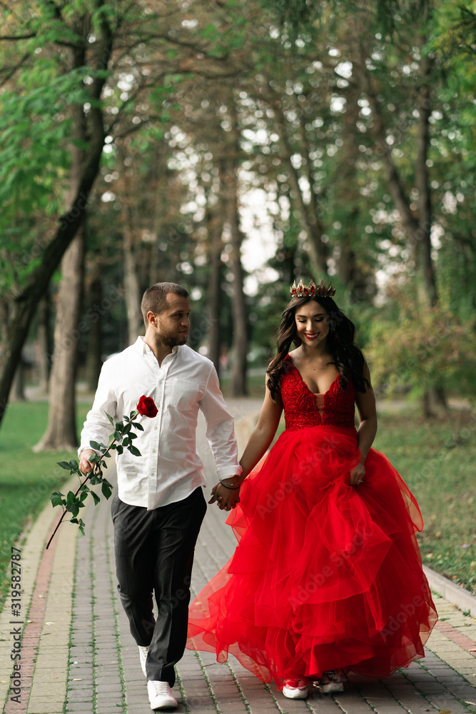 Beautiful romantic couple. Attractive young woman in red dress and crown with handsome man in white shirt with red rose walking on the street.Happy Saint Valentine's Day. Pregnant and wedding concept.