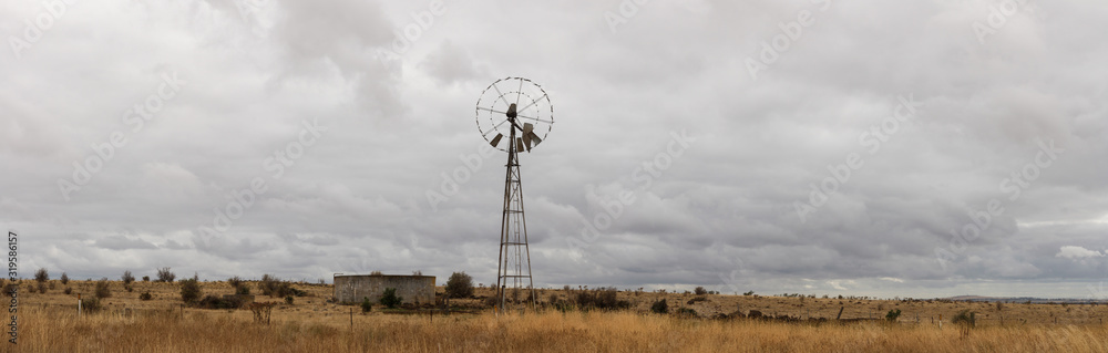 typical farming scene of an old tin wind powered water pump standing tall in an open field full of dry, drought affected grassland, on a cloudy overcast day near Melbourne, Victoria, Australia