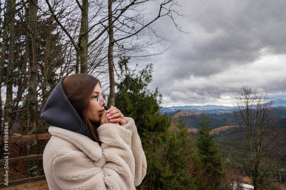 Side view portrait of the beautiful brunette girl in beige coat and blue hood holds her arms near her face, beautiful mountains view on the background Side view portrait of the beautiful brunette girl in beige coat and blue hood holds her arms near her face, beautiful mountains view on the background