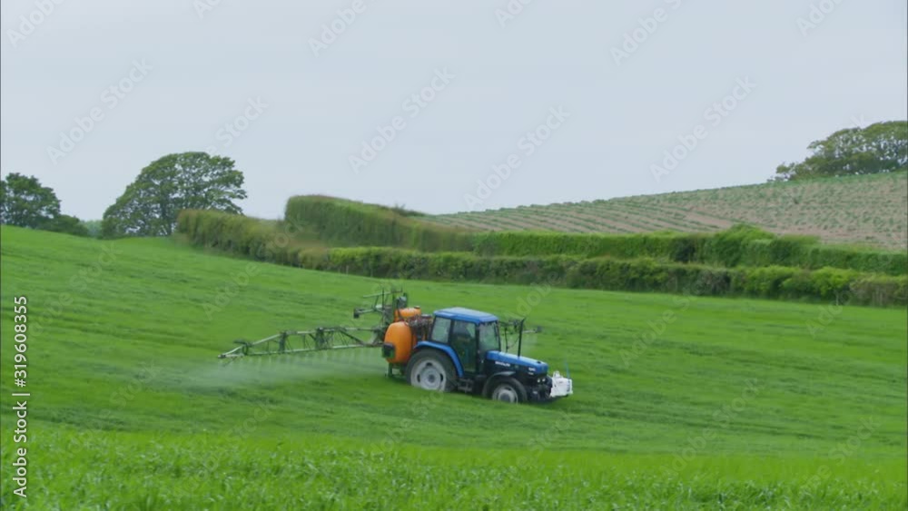 Wide low angle panning shot of a green tractor farm with orange boom sprayer spraying a lush green grass fiend, UK Wide low angle panning shot of a green tractor farm with orange boom sprayer spraying a lush green grass fiend, UK