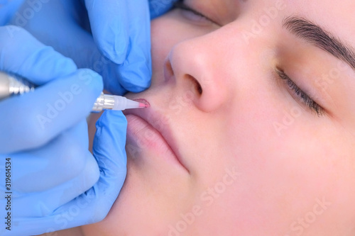 Cosmetologist is applying permanent makeup on lips using tattoo machine in beauty clinic. Beautician hands and patient face closeup. Lips microblading procedure for girl. Beauty industry concept.