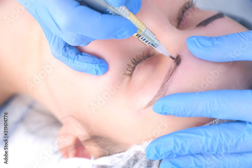 Cosmetologist making eyebrows microblading procedure in beauty salon for girl using tattoo apparatus  eyebrow closeup. Beautician in gloves is doing permanent makeup to woman. Beauty industry concept.