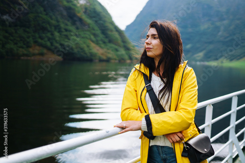 The girl tourist on a pleasure boat on the fjord enjoys the picturesque mountains and lakes of Norway. Young woman posing against the backdrop of the mountains. Travelling, lifestyle, adventure. © maxbelchenko