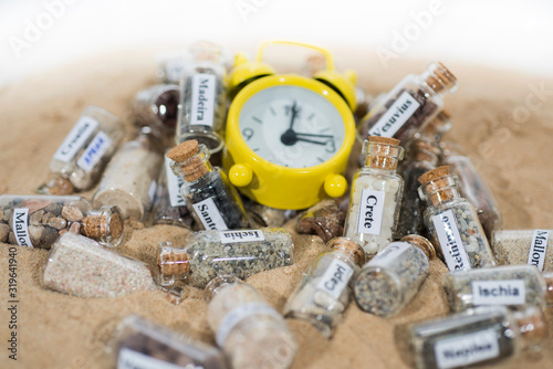 Glass test-tube with sand of different summer vacation destinations. Located in sand with clock. Vacation time abstract photo.