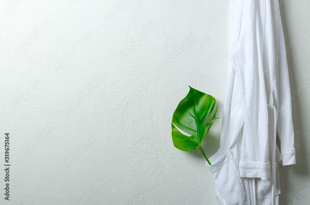 Fresh white bathrobe hanging on the hook and green leaf in the pocket as a decoration.Concept of leisure spa.Empty space for text Fresh white bathrobe hanging on the hook and green leaf in the pocket as a decoration.Concept of leisure spa.Empty space for text
