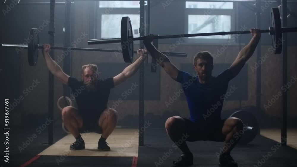 Two strong man doing weightlifting training together at the gym. Medium shot slow motion pan men exercising in gym with exercise equipments: Overhead squats, plate lunges, medicine ball, olympic rings Two strong man doing weightlifting training together at the gym. Medium shot slow motion pan men exercising in gym with exercise equipments: Overhead squats, plate lunges, medicine ball, olympic rings
